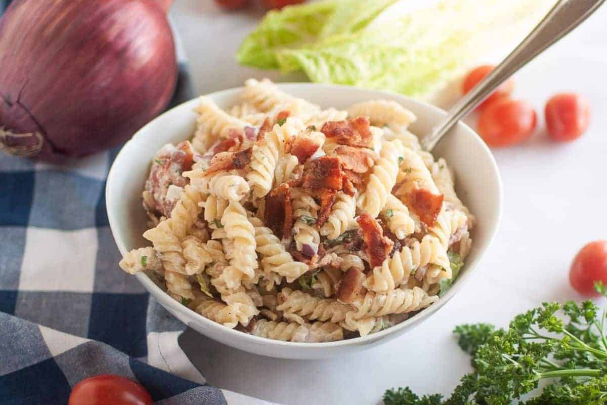 A white bowl filled with BLT pasta salad, featuring spiral pasta, creamy dressing, pieces of bacon, and other visible ingredients, is shown with a spoon. Red onions, lettuce, cherry tomatoes, and parsley are in the background.