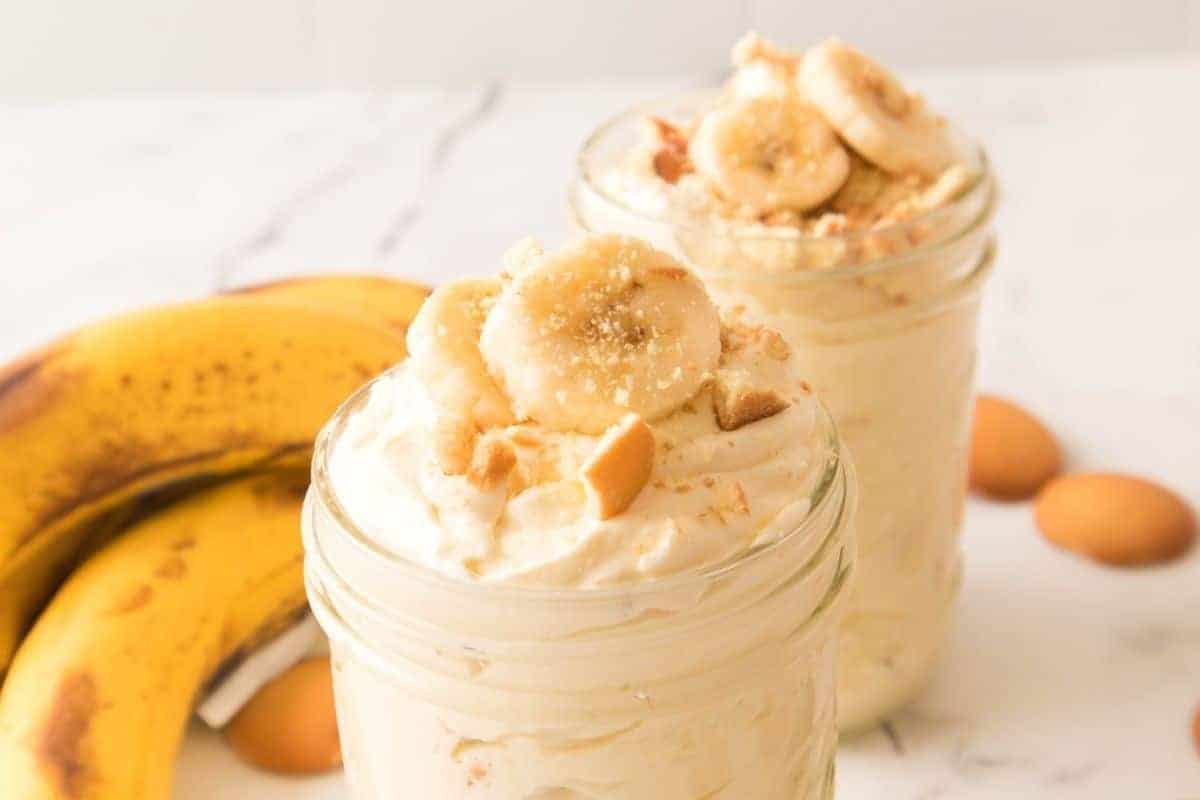Two jars of banana pudding topped with banana slices and crushed cookies are in the foreground, with bananas and vanilla wafers in the background on a white surface.