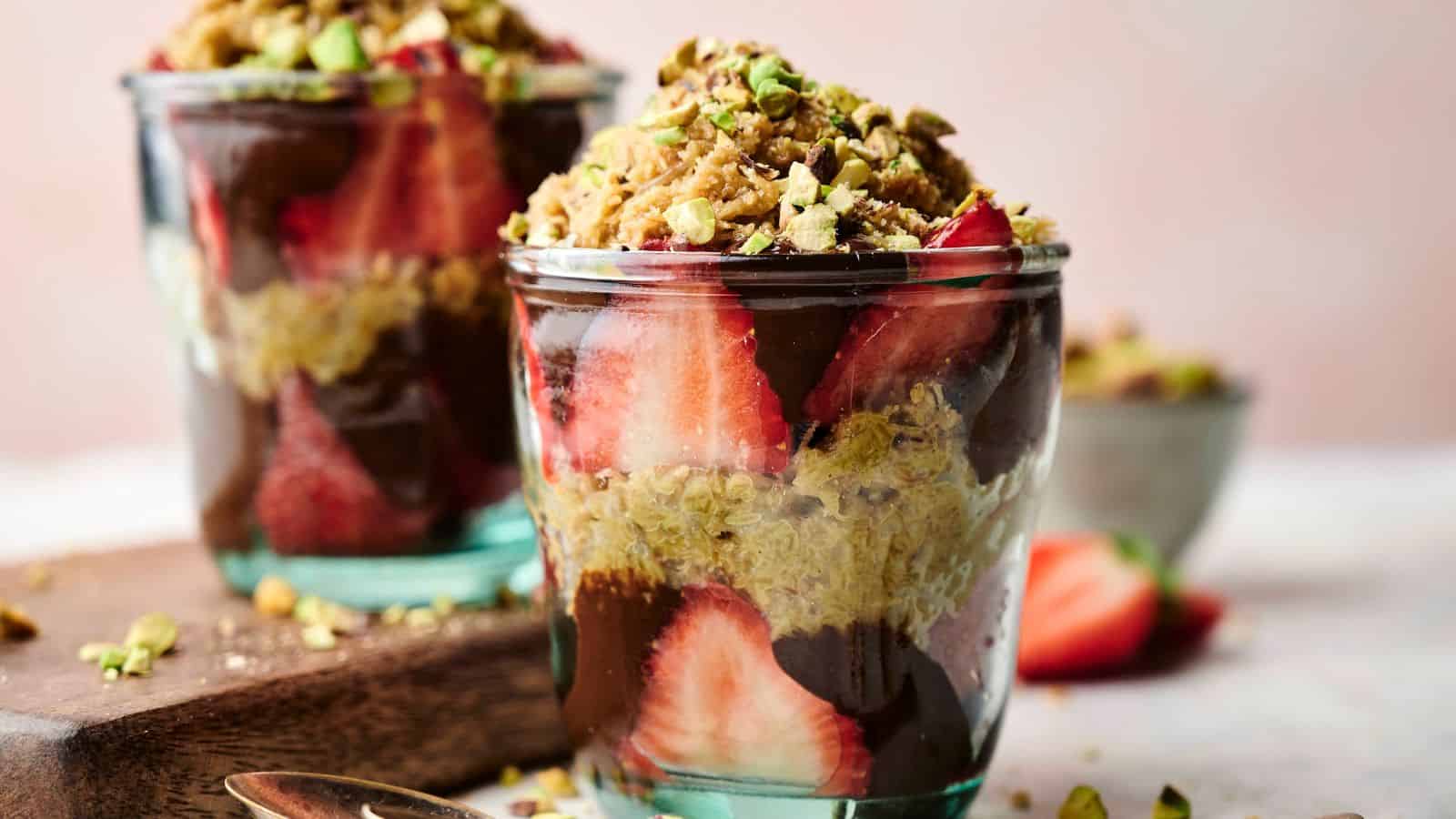 Two glass cups filled with layers of sliced strawberries, chocolate, granola, and chopped pistachios, with a blurred bowl of nuts in the background.