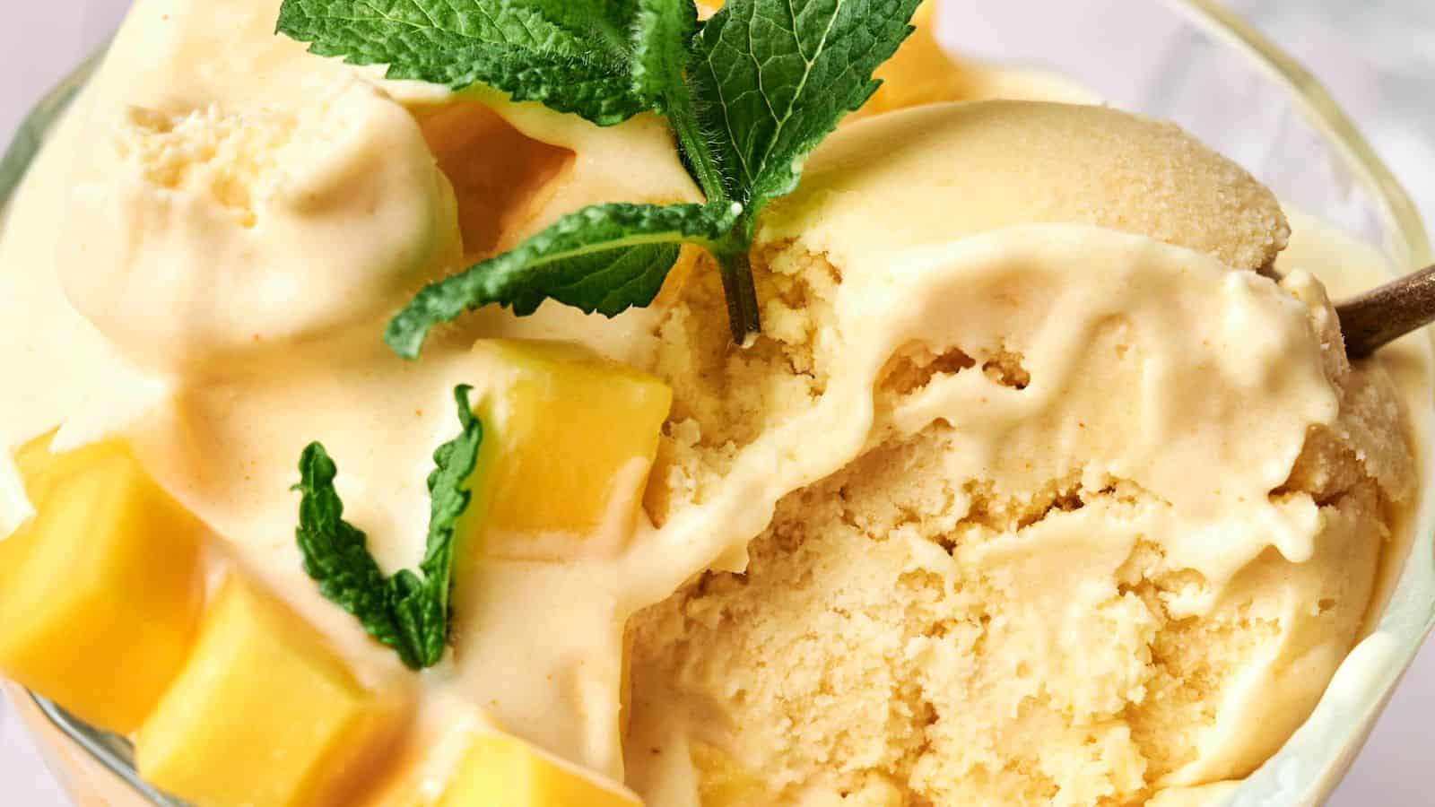 A bowl of creamy mango ice cream is garnished with fresh mango cubes and mint leaves.