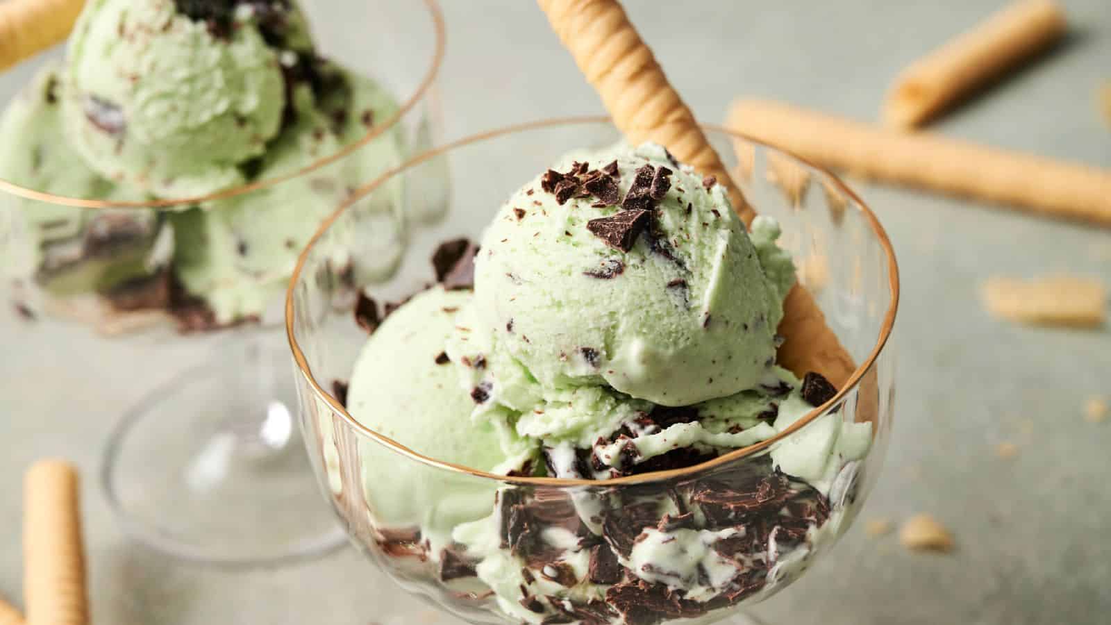 A glass dessert cup filled with mint chocolate chip ice cream, topped with chocolate pieces and served with rolled wafer cookies.