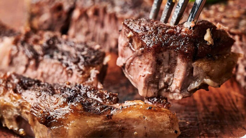 Close-up of cooked beef slices on a wooden board, with a fork holding one piece. The meat appears juicy with a seasoned crust.