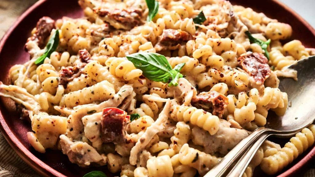 A plate of creamy rotini pasta with shredded chicken, sun-dried tomatoes, fresh basil, and cracked black pepper—just as effortless as the best salads for lazy weeknights, served with a spoon.