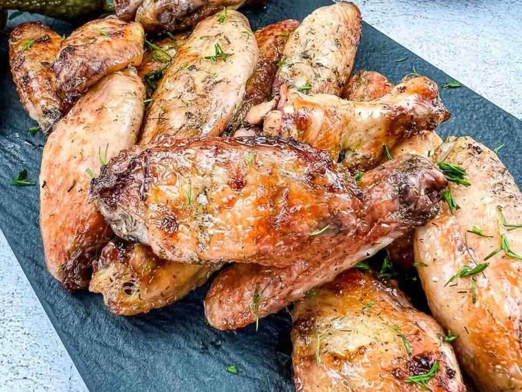 A pile of cooked chicken wings with a golden-brown, crispy skin garnished with herbs, served on a dark rectangular slate platter.