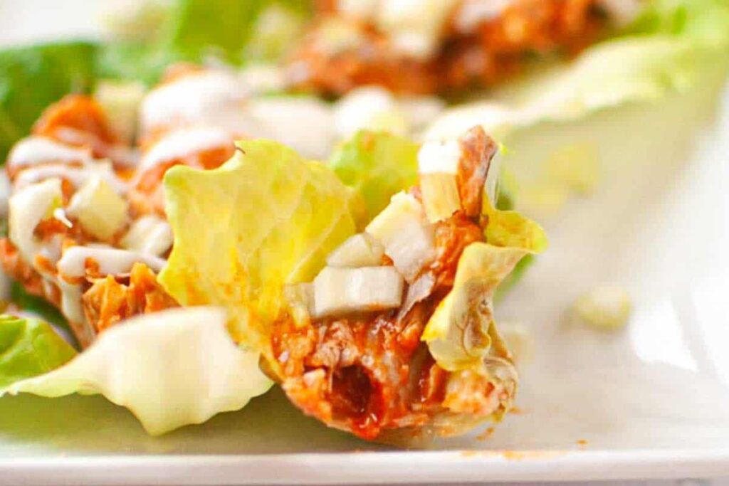 Close-up of a lettuce wrap filled with shredded meat, diced onions, and a drizzle of white sauce on a white plate.