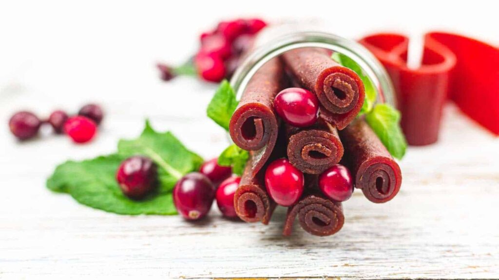 Rolled cranberry fruit leather strips and fresh cranberries are arranged on a white wooden surface, with a glass jar and green leaves in the background.