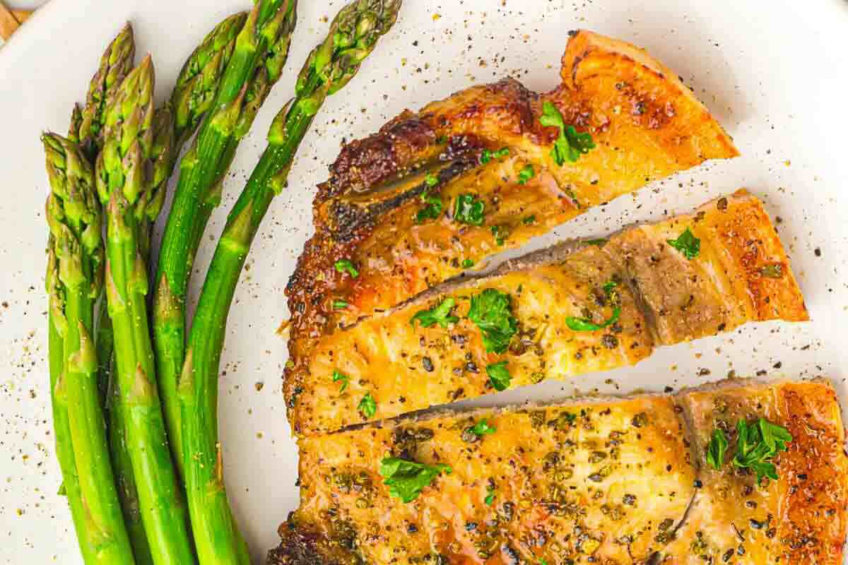 Cooked crispy pork chop garnished with herbs, served alongside steamed asparagus on a white plate.