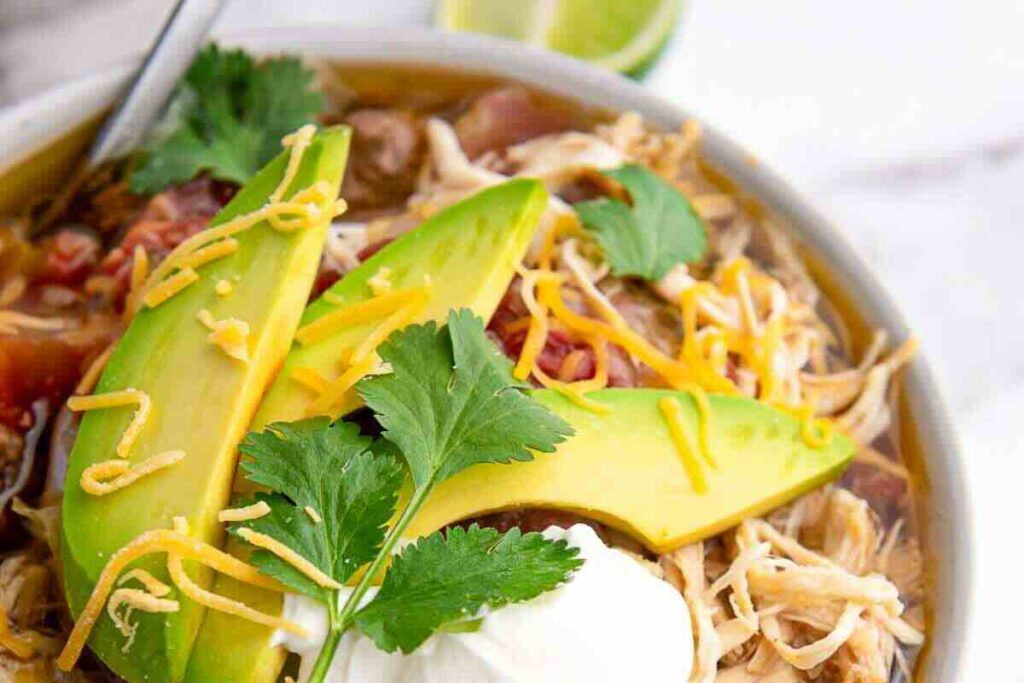 A bowl of shredded chicken soup topped with sliced avocado, shredded cheese, cilantro, and a dollop of sour cream.
