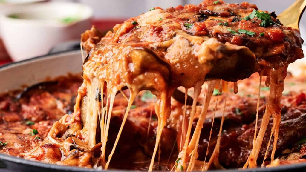 A close-up of a cheesy, baked lasagna slice being lifted from a dish, with melted cheese stretching and tomato sauce visible.