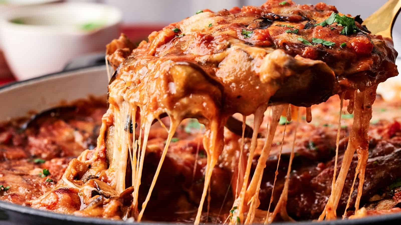 A slice of cheesy eggplant lasagna with tomato sauce is being lifted from a pan, showing stretchy melted cheese. A bowl is blurred in the background.
