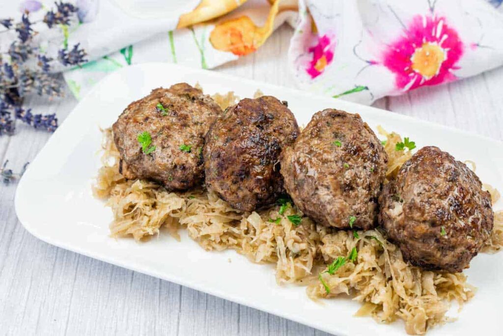 Four German meat patties are served on a bed of sauerkraut, garnished with parsley, on a rectangular white plate with a floral napkin in the background.
