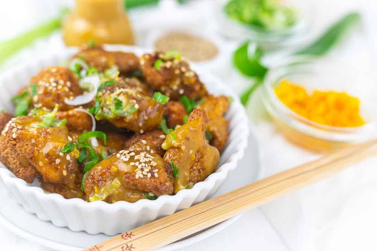 A white dish filled with orange chicken topped with sesame seeds and green onions, surrounded by small bowls of toppings and a pair of chopsticks.