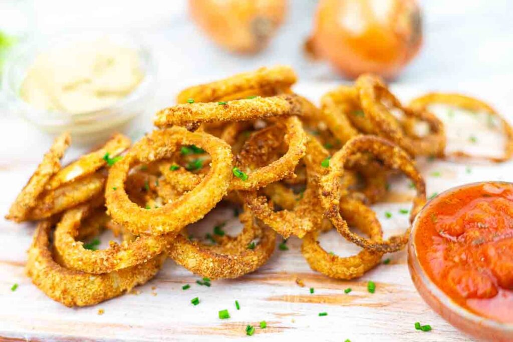 A pile of golden-brown onion rings garnished with chopped chives, served on a wooden surface with red and yellow dipping sauces in small bowls.