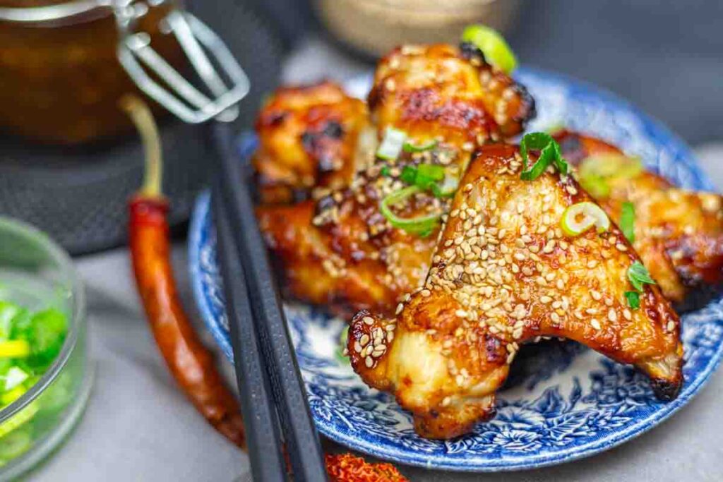 Grilled chicken wings topped with sesame seeds and chopped green onions are served on a blue patterned plate with chopsticks.
