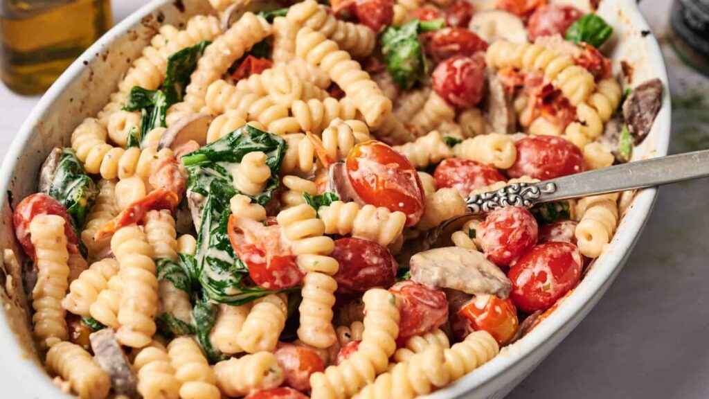 A serving dish filled with curly pasta, cherry tomatoes, spinach, mushrooms, and a creamy sauce, with a spoon resting inside.
