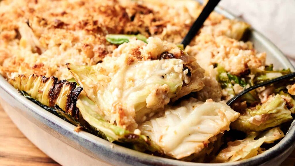 A baked casserole featuring layers of cabbage and a creamy, breadcrumb topping, with serving utensils in the dish.