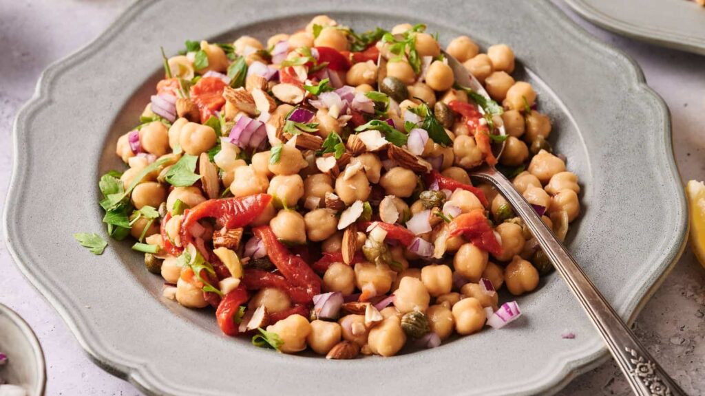 A plate of chickpea salad with chopped red onion, roasted red peppers, capers, herbs, and almonds, served with a spoon.