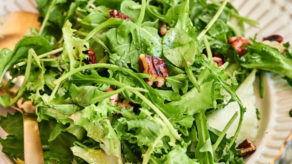 A close-up of a fresh green salad with leafy greens, pecans, and a light vinaigrette on a white plate.
