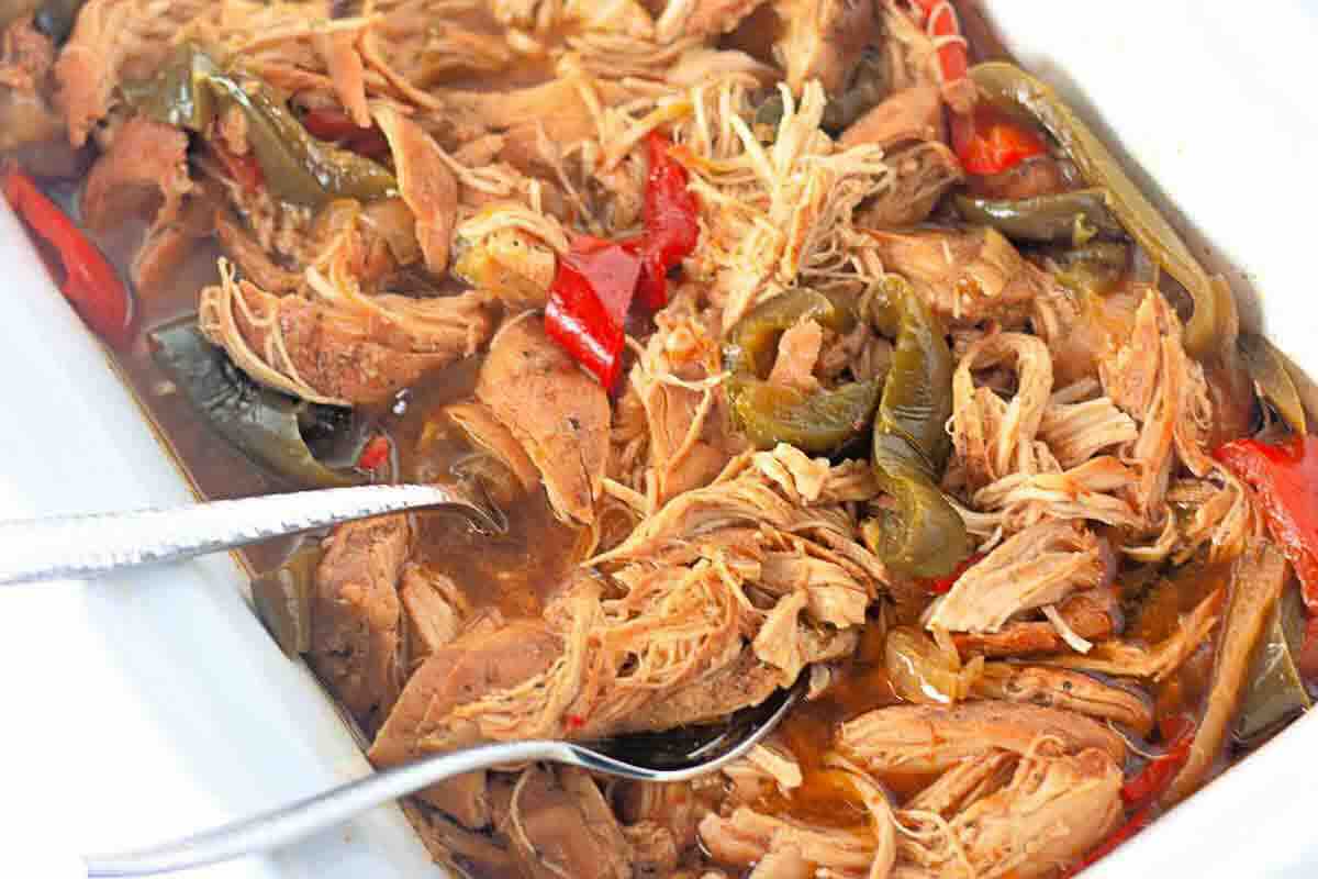 Shredded cooked chicken with red and green bell peppers in a sauce, served in a white dish with metal tongs and a spoon.