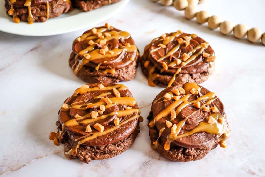 Four chocolate cookies topped with chocolate frosting, caramel drizzle, and chopped nuts are arranged on a marble surface.