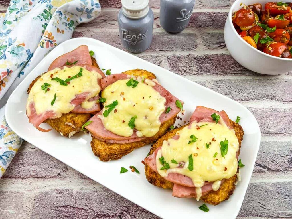 Three pieces of breaded chicken breasts topped with ham, melted cheese, and chopped herbs are arranged on a white plate, with a bowl of tomato salad and salt and pepper shakers nearby.