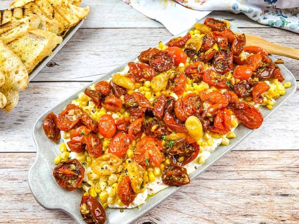 A rectangular platter topped with roasted cherry tomatoes, corn, and cheese, served on a wooden table next to sliced grilled bread.