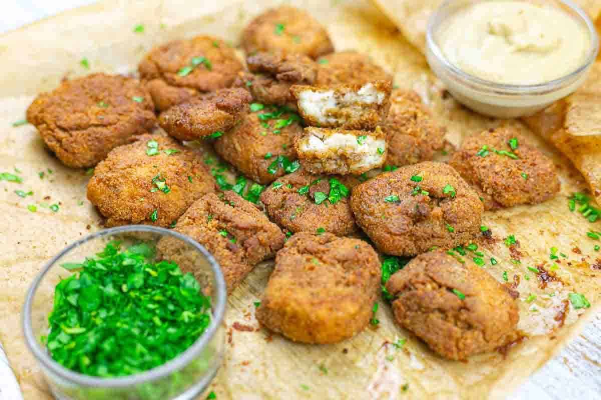 A batch of breaded and fried nuggets is arranged on parchment paper, garnished with chopped herbs, with two small bowls of dipping sauces nearby.