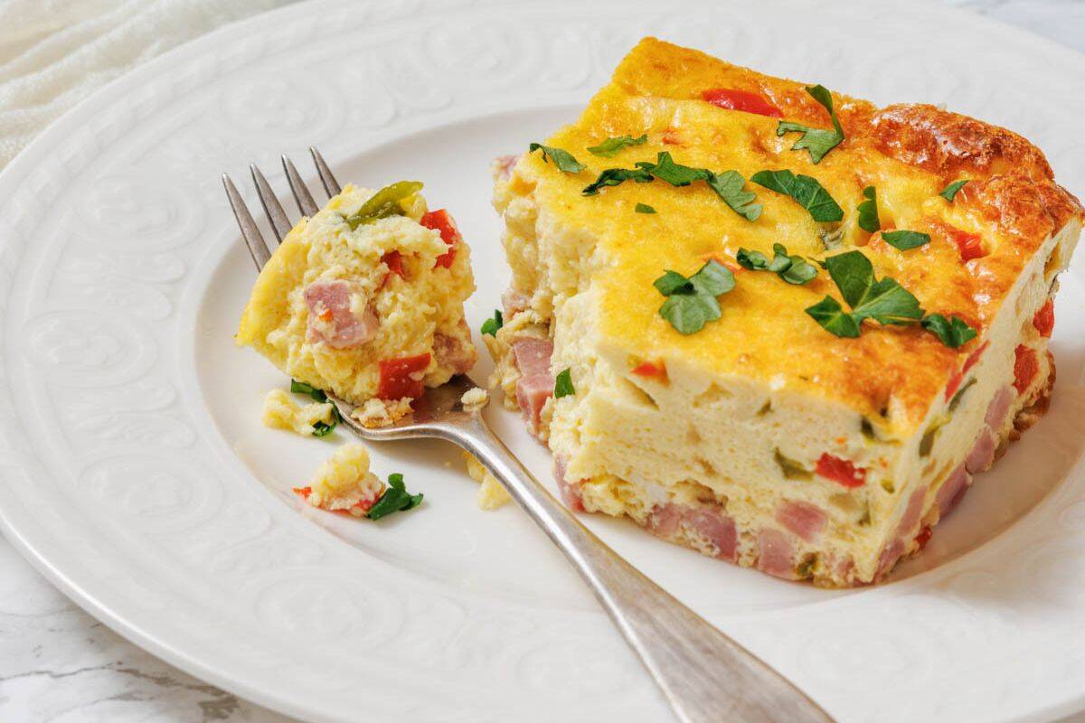 A slice of baked Western omelet with ham, red peppers, and herbs is served on a white plate with a fork holding a bite-sized piece.