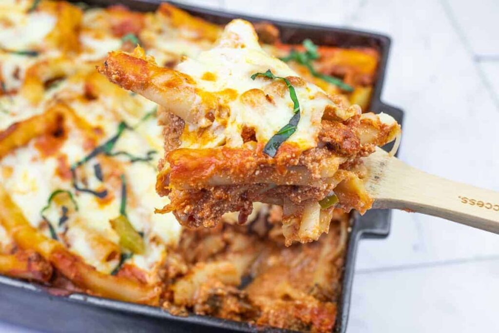 A close-up of a wooden spoon lifting a serving of baked ziti with melted cheese and tomato sauce from a casserole dish.