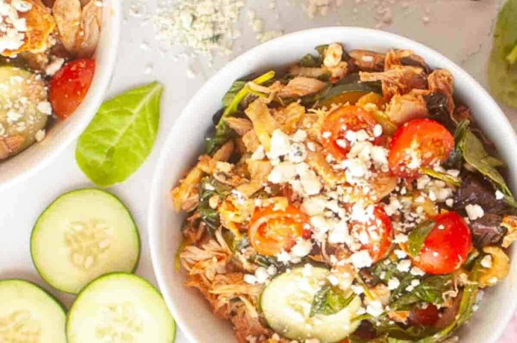 A bowl of salad with shredded chicken, cherry tomatoes, cucumbers, leafy greens, and crumbled cheese, with cucumber slices on the side.