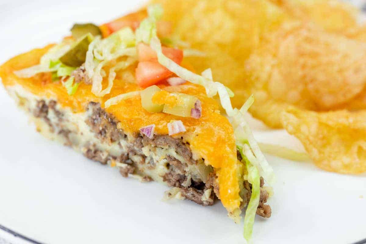 A Cheeseburger Pie topped with shredded lettuce, diced tomatoes, pickles, and chopped red onions in a white pie dish on a light wooden surface.
