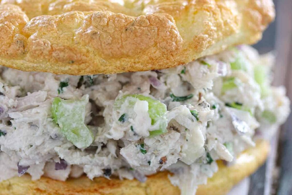 Close-up of a chicken salad sandwich with celery and herbs on a fluffy, golden-brown bread roll.