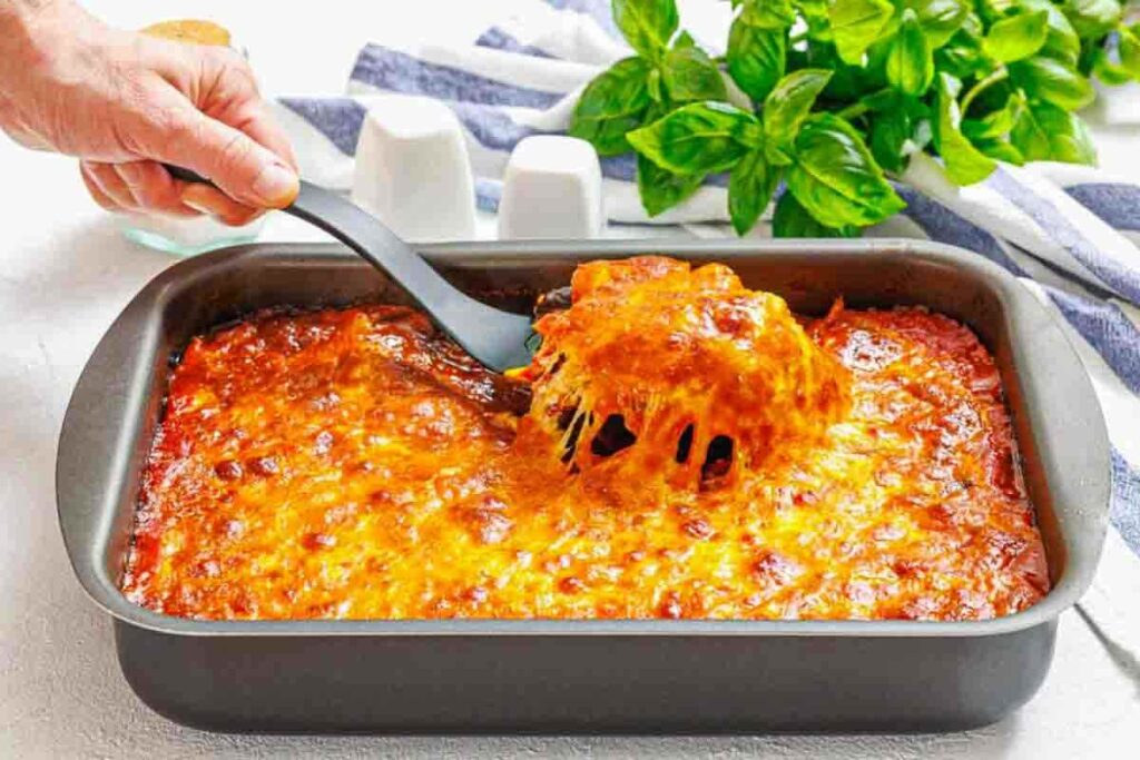 A hand serves a portion of cheesy baked lasagna from a rectangular pan, with melted cheese stretching, placed on a white surface near fresh basil.
