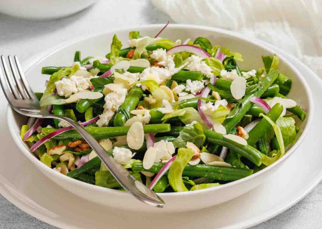 A bowl of green bean salad with feta cheese, sliced red onions, almonds, and mixed greens, served with a fork on a white plate.