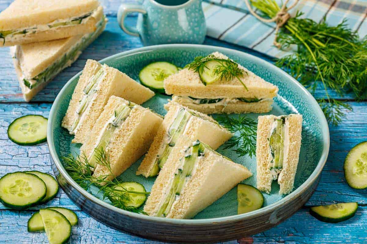 A plate of triangular cucumber sandwiches on white bread, garnished with fresh dill and cucumber slices, set on a blue wooden table.