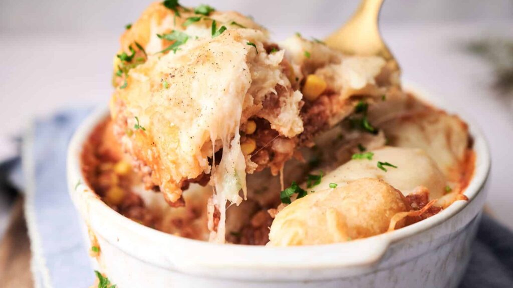 A bowl of shepherd's pie with a spoon lifting a portion, showing layers of meat and mashed potatoes, garnished with parsley.