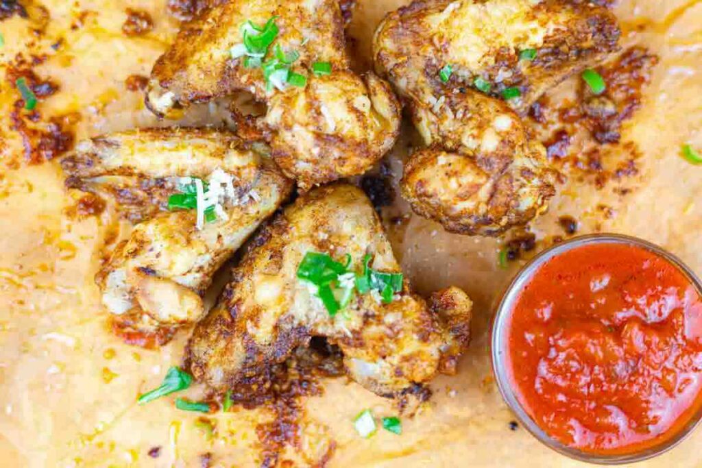 Five seasoned baked chicken wings on parchment paper, garnished with chopped green herbs, served with a small cup of red dipping sauce.
