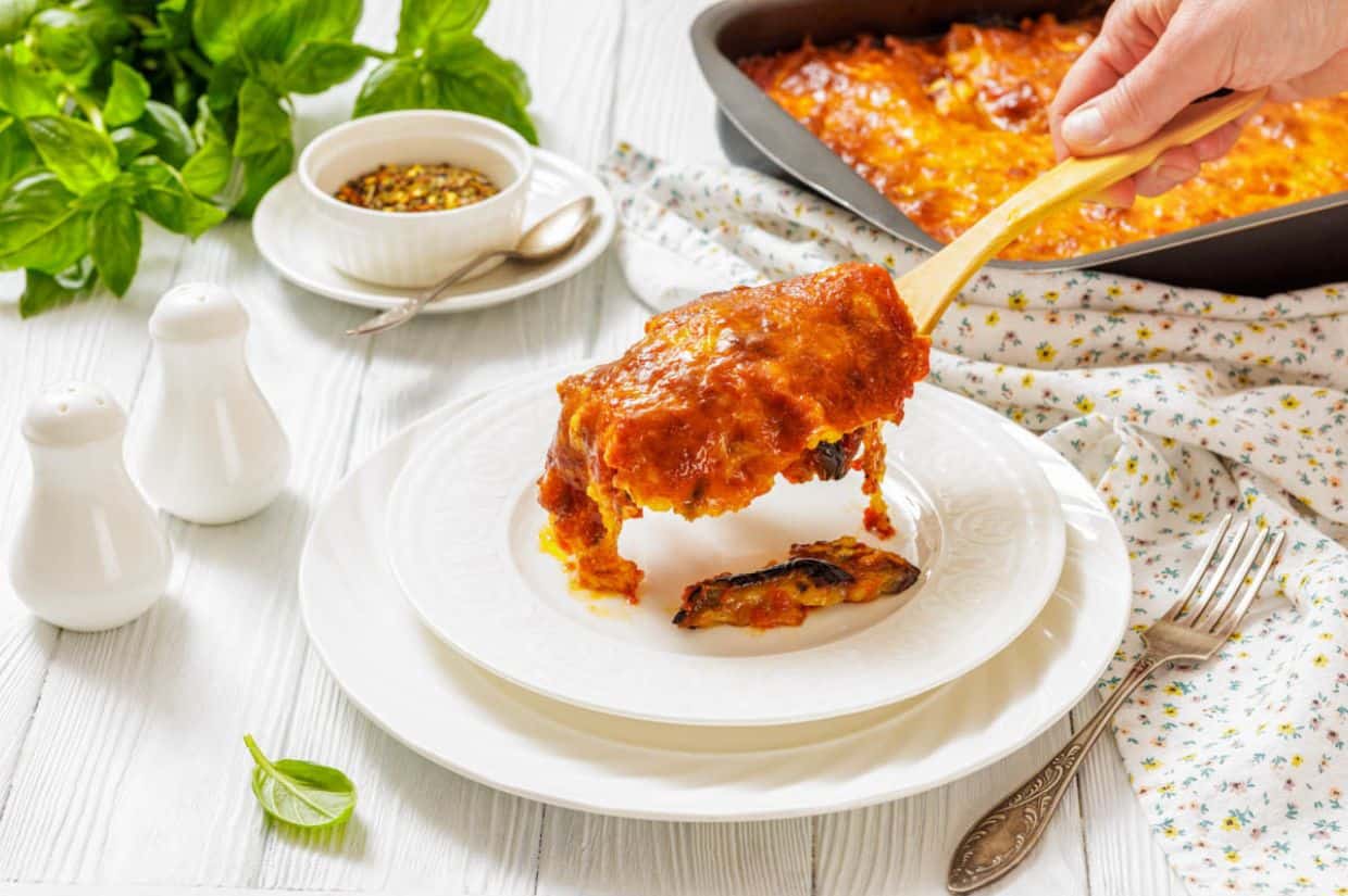 A hand serves a slice of Eggplant Parmigiana onto a white plate, with a casserole dish, herbs, and condiments visible on a white table.