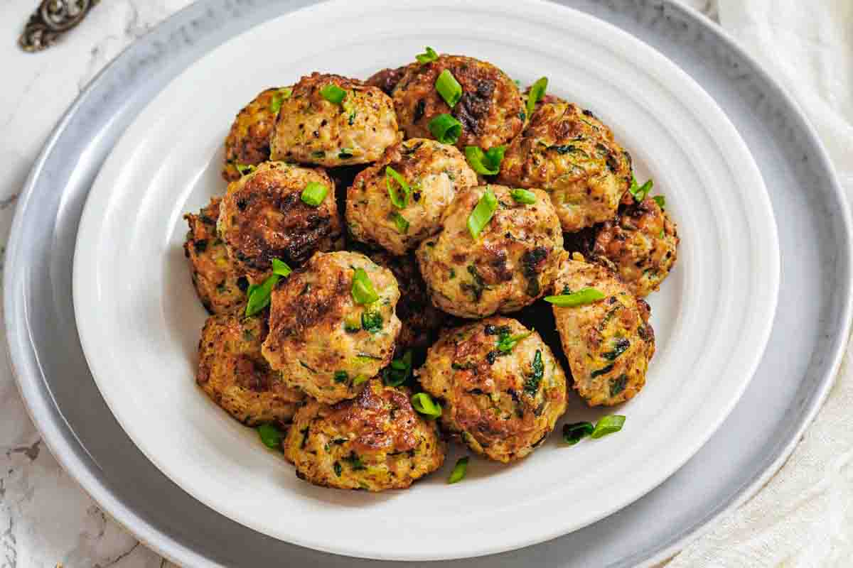 A white plate filled with cooked zucchini fritters garnished with chopped green onions, placed on a larger gray plate.