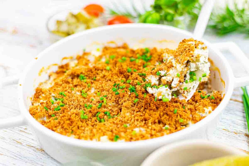 A white casserole dish filled with a baked creamy mixture topped with golden breadcrumbs and chopped green herbs, with a serving spoon lifting a portion.