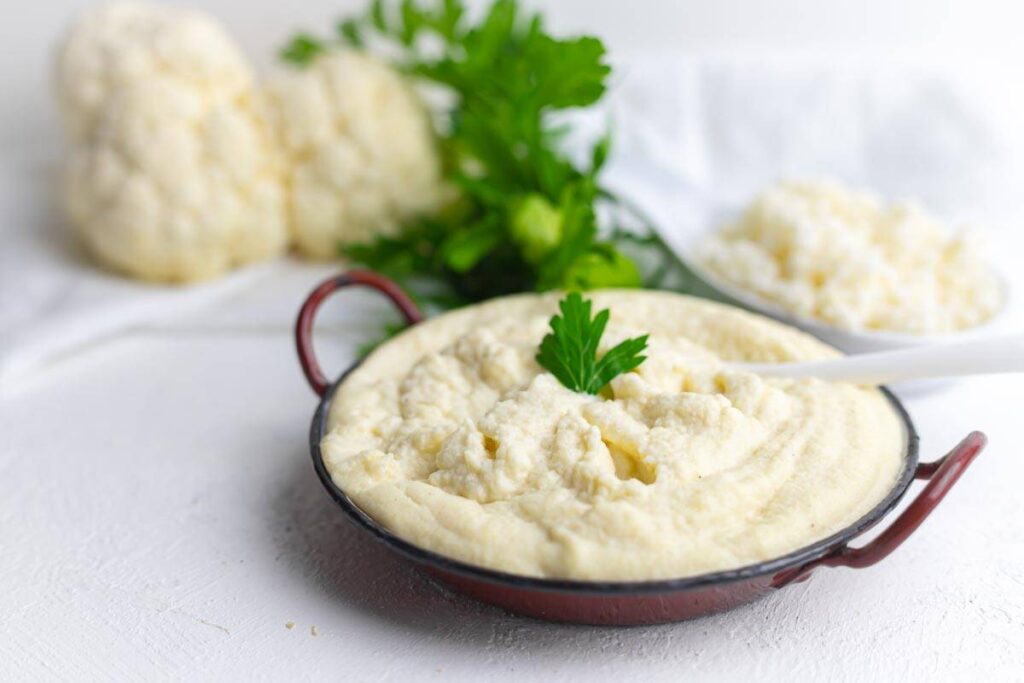A bowl of creamy cauliflower mash garnished with a parsley leaf, with fresh cauliflower and chopped parsley in the background.