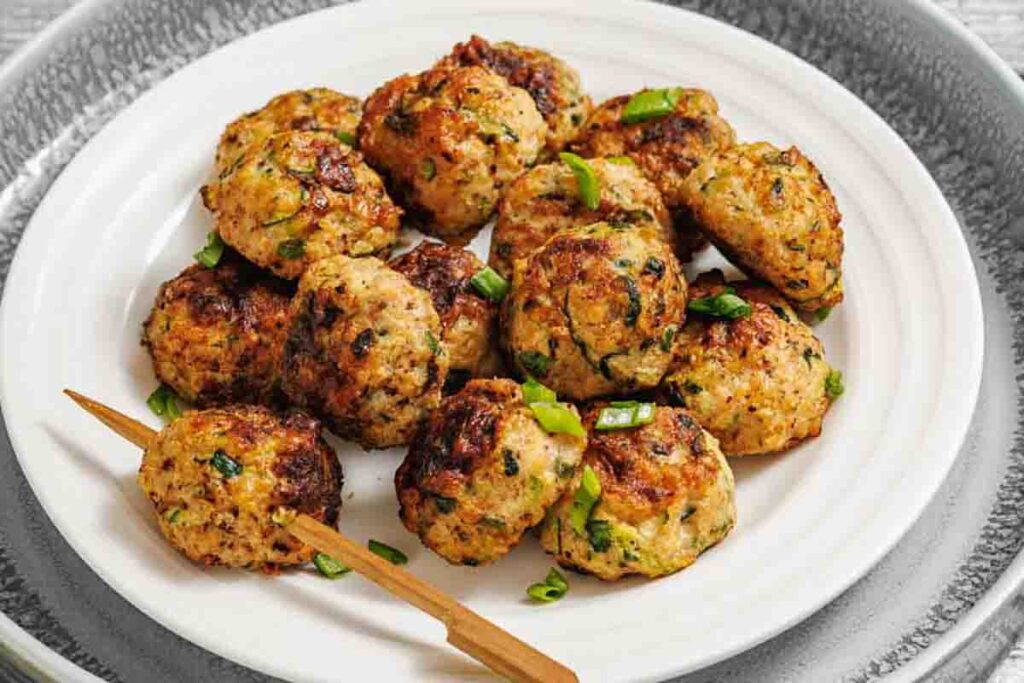 A white plate with several browned meatballs garnished with chopped green onions, with a wooden pick resting on the plate.