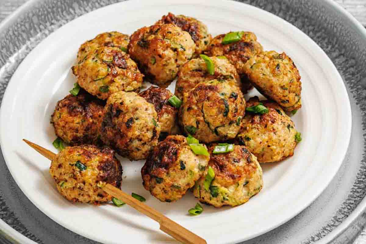 A white plate with several zucchini patties garnished with chopped green onions, and a wooden skewer resting on the edge.