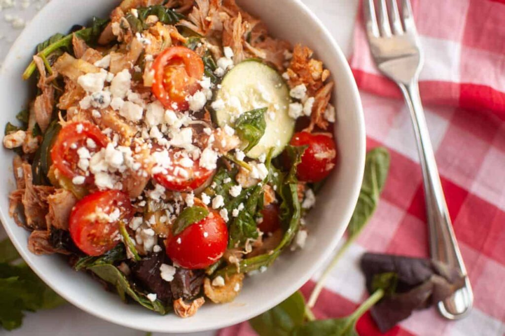 A bowl of Buffalo Chicken salad with shredded meat, cherry tomatoes, spinach, sliced cucumber, and crumbled cheese, next to a red-and-white checkered napkin and a fork.