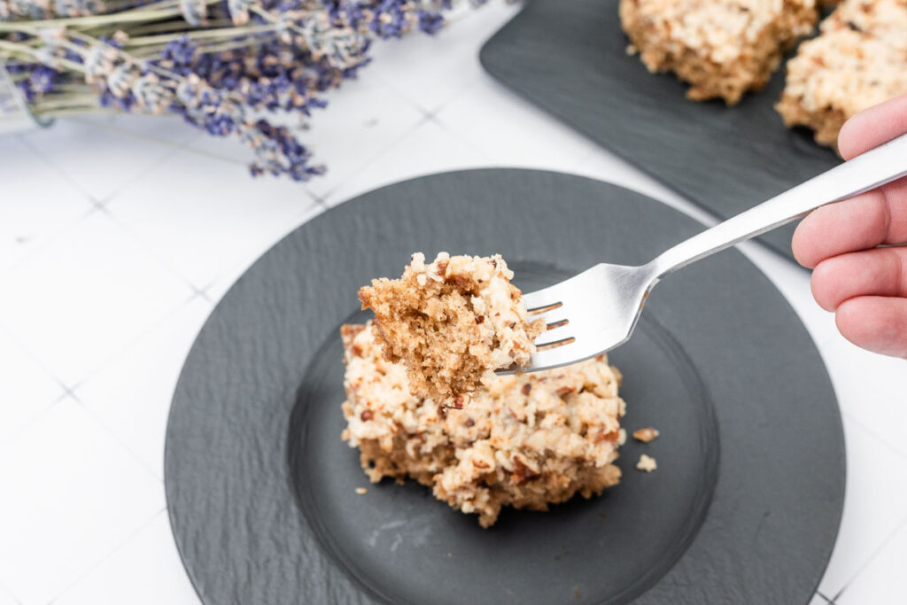 A fork holds a bite of Old Fashioned Lazy Daisy Oatmeal Cake above a black plate with more cake pieces, with dried lavender in the background.