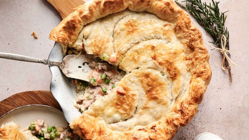 A golden-brown chicken pot pie with a slice removed, revealing a filling of diced vegetables and chicken. A serving spatula rests in the pie dish, and rosemary is placed nearby.