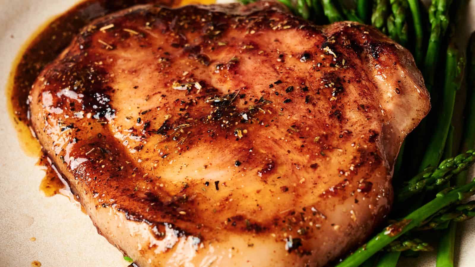 A cooked pork chop with brown sauce is served on a white plate alongside steamed asparagus spears.