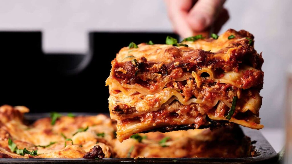 A person lifts a slice of lasagna from a pan, revealing layers of pasta, meat, cheese, and tomato sauce. The lasagna is garnished with fresh herbs.