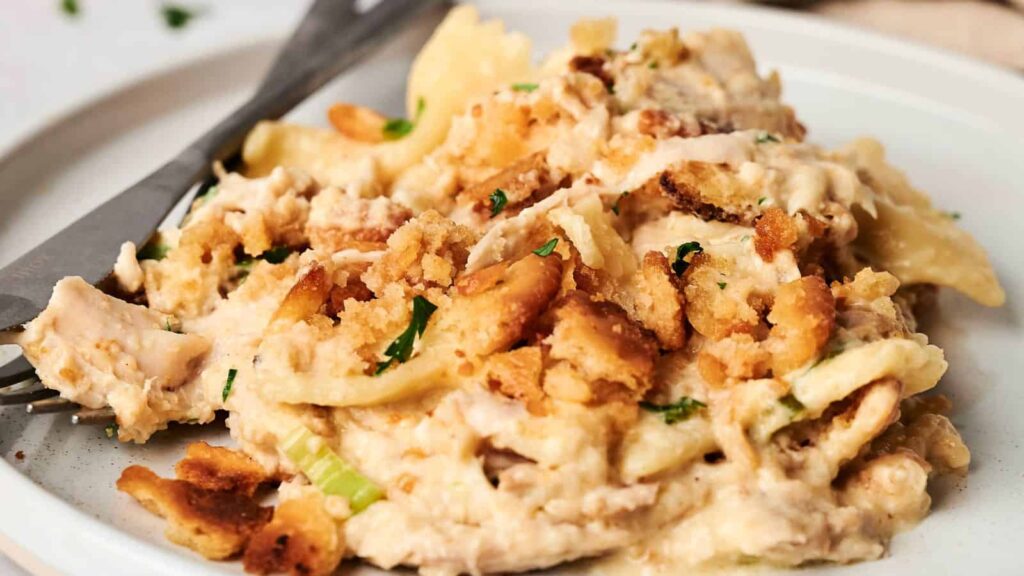 A plate of creamy chicken casserole with bowtie pasta and a crumb topping. A fork and knife are placed on the side of the plate.
