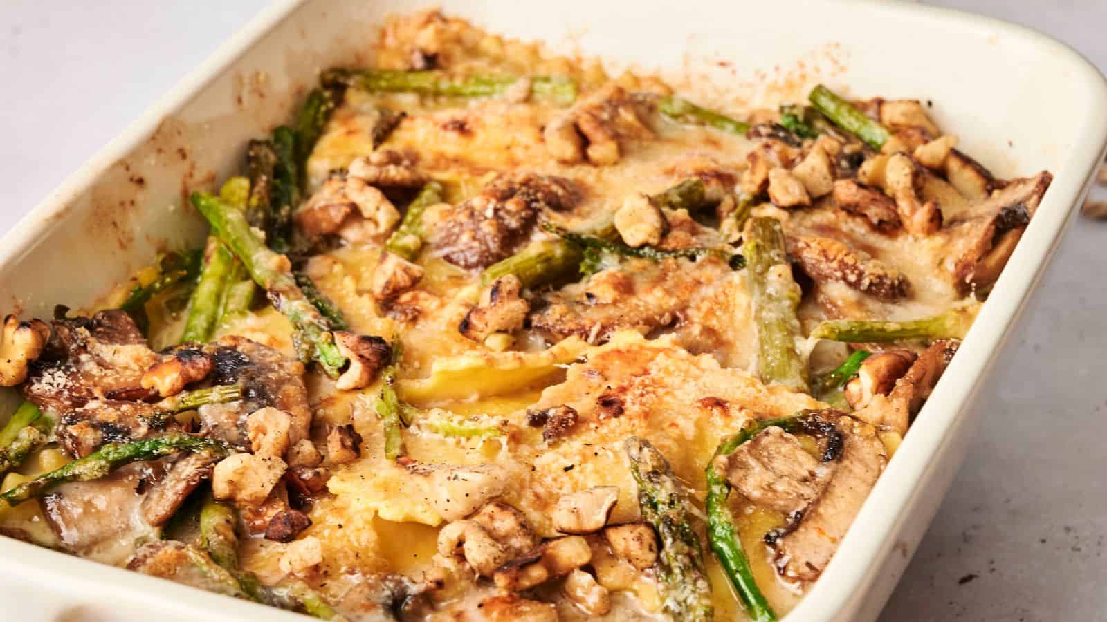 Close-up of a baked dish with ravioli, mushrooms, asparagus, and melted cheese on top.